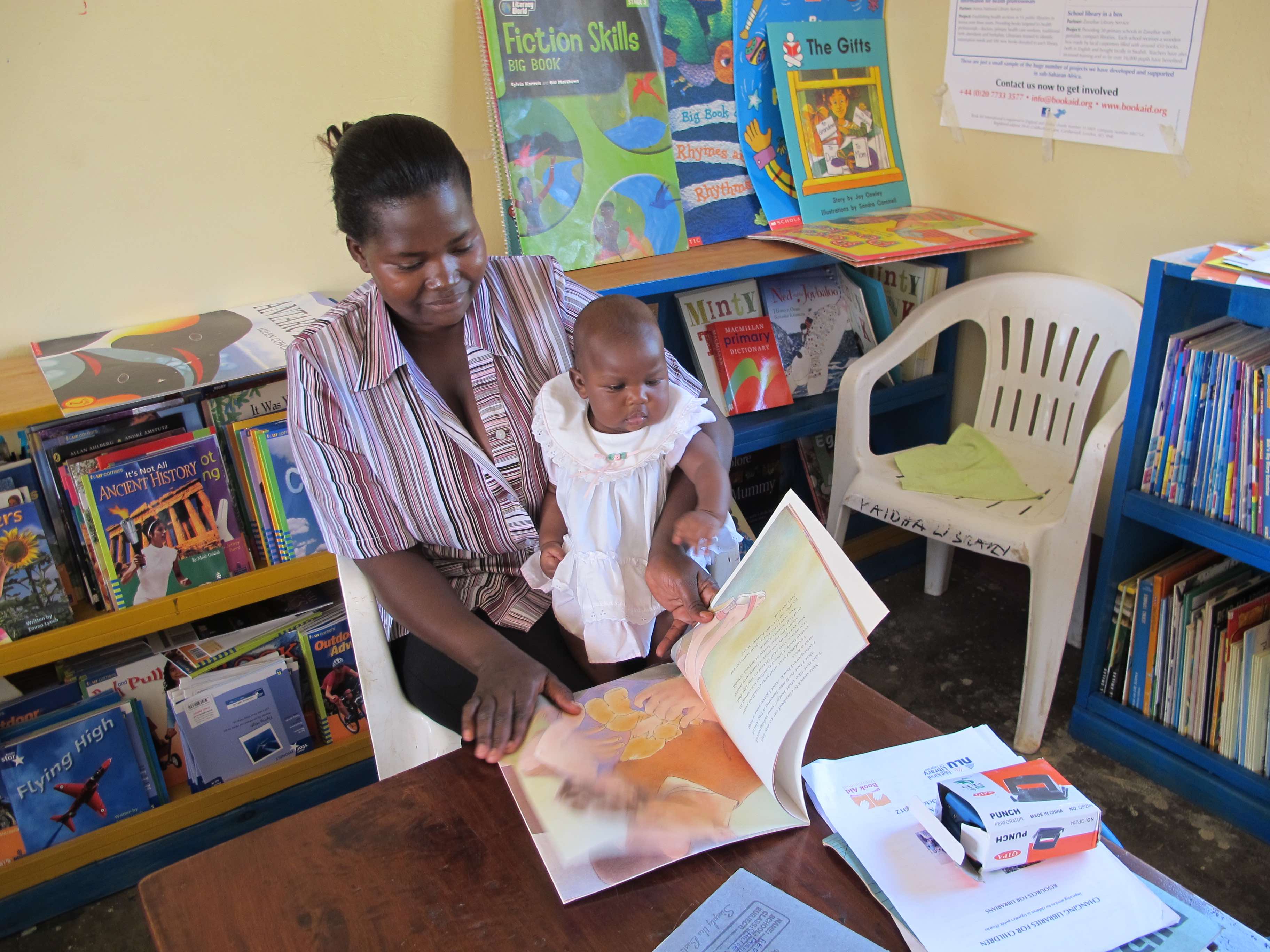 A toddler sits on the lap of an adult in the children’s reading room at the National Library of Uganda. The adult is paging through a picture book, while the child looks at the pictures. The library shelves behind the adult and against the walls are stacked with colourful picture books.